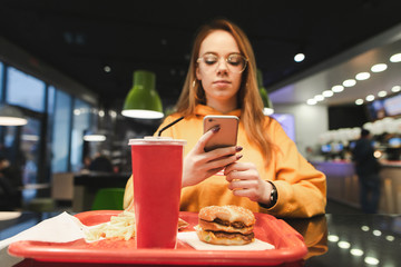 Girl uses a smartphone and dishes with a fast food. Attractive, stylish girl wearing glasses and gourmet clothes sitting at the fast food restaurant table with a tray of food and using a smartphone.