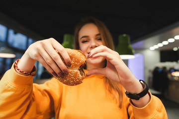 Attractive girl in orange clothing holds in the hand a great tasty burger and shows a sign of the heart. Girl loves burgers. Love for fast food. Copyspace - obrazy, fototapety, plakaty