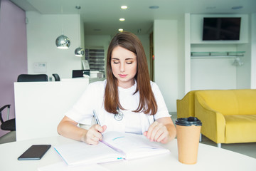 Portrait of young female doctor making notes in notebook while sitting behind the table with phone and coffee inside of medical clinic. Student on practice. Healthcare and medicine. Copy space.