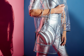 partial view of model in metallic bodysuit and raincoat posing with silver bananas for fashion shoot on pink and blue background