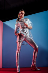 fashionable young woman in metallic bodysuit and raincoat posing with silver bananas on blue background