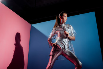 beautiful fashionable woman in metallic bodysuit and raincoat posing with silver bananas on pink and blue background