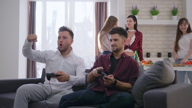 weekend, guys play video games on the consoles sitting on sofa on backgrounf of company of friends enjoy rest indoor in modern apartment