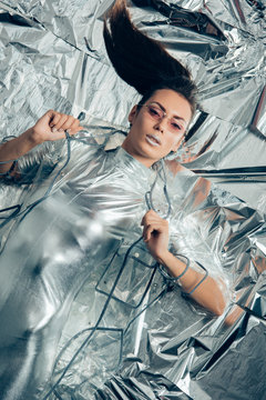 top view of brunette fashionable girl posing in silver bodysuit and raincoat on metallic background
