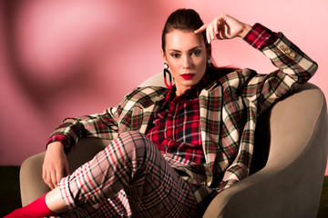 fashionable woman in checkered suit posing in armchair on pink background