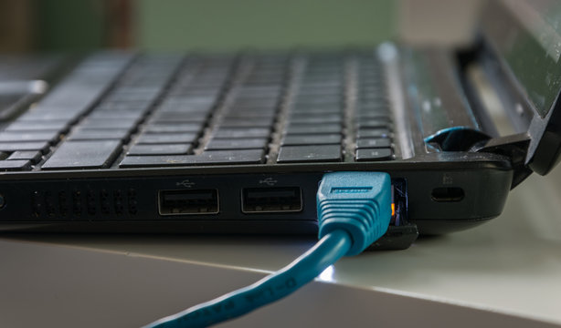  Blue ethernet cable connected to a black laptop on a white table. The lights near the cable head are on.