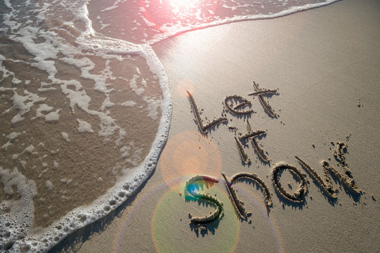 Let it Snow winter travel message handwritten in the smooth sand of a tropical beach with sunny lens flare