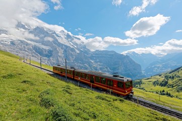 Fototapeta na wymiar July, 6, 2018, A cog wheel train travels on famous Jungfrau Railway from Kleine Scheidegg to Jungfraujoch station ( top of Europe ) on a green grassy hillside, in Berner Oberland, Switzerland 