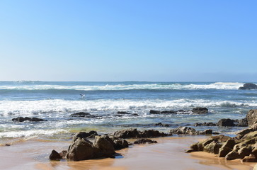 Fototapeta na wymiar Wild beach with rocks and big waves with foam and mist. Blue sky, sunny day, Galicia, Spain.