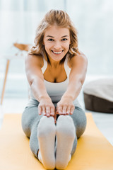 smiling woman doing stretching exercise on yellow fitness mat and looking at camera at home