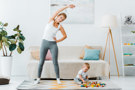 smiling woman doing exercise and looking at camera and toddler boy playing with multicolored cubes on carpet in living room