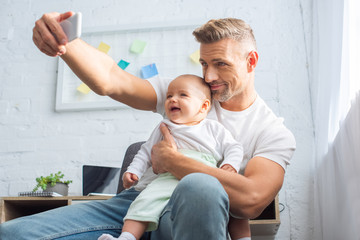 Fototapeta na wymiar happy father sitting on chair, holding baby daughter and taking selfie on smartphone at home