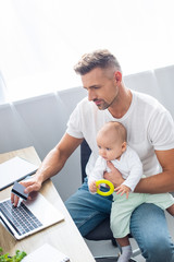 handsome father sitting at computer desk, doing online shopping with credit card and holding baby daughter at home