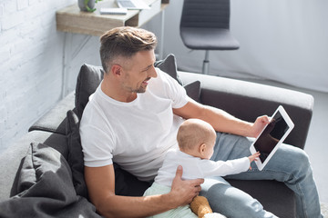 smiling father sitting on couch with adorable baby daughter and using digital tablet with blank screen at home