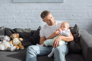 smiling father sitting on couch and feeding baby daughter in living room