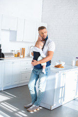 serious father holding infant daughter in baby carrier and talking on smartphone