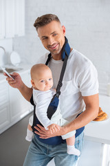 cheerful father using smartphone and holding infant daughter in baby carrier