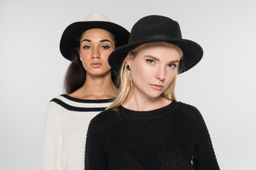 portrait of beautiful multicultural women in black and white clothes and hats looking at camera isolated on white