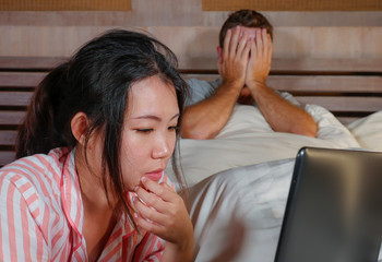 frustrated husband moody in bed ignored by his workaholic Asian wife or internet social media addict girlfriend using laptop in bed ignoring the man in couple internet conflict