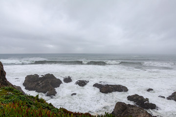 The aftermath of a recent storm gives overcast sky, heavy surf, big waves crashing on the rocks near the shoreline.