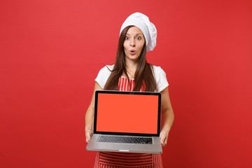 Housewife female chef cook or baker in striped apron white t-shirt toque chefs hat isolated on red wall background. Woman hold laptop pc blank screen for promotional content Mock up copy space concept