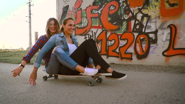 two beautiful and young girlfriends having fun with skateboard outdoor, in the evening sun