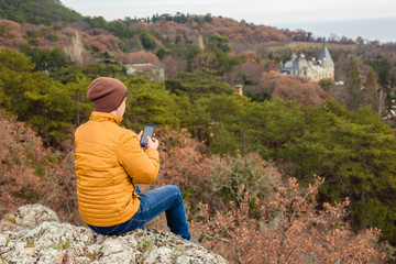 Naklejka premium The traveler of the yellow jacket looks with the phone in his hands from the mountain to the pine forest. Tourism, active recreation.