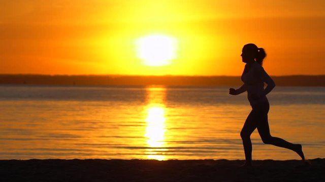 Silhouette of sporty woman running in sea ocean beach at sunset. Female jogger jogging in nature. Athletic girl running outdoor. Healthy fitness wellness sport concept. Sun rays slow-motion