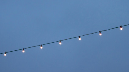 Blue sky background. Electric garland for lighting with white light bulbs against the background of...