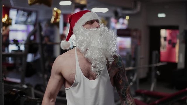 Santa Claus in the gym is grimacing and showing biceps.