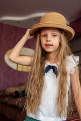 Cute blond girl in school uniform are posing in front of camera with straw hat in her hands inside house. Long hair. Child happy holidays.