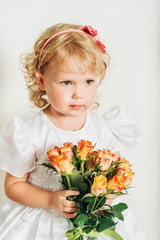 Studio shot of adorable 3 year old toddler girl wearing white occasion dress, holding bouquet of yellow orange roses