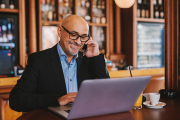 Bearded senior in suit using smart phone and laptop while sitting in cafeteria. On the table laptop, juice and coffee.