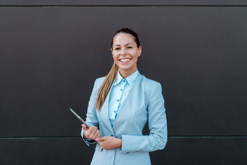 Smiling beautiful brunette in suit holding tablet and looking at camera while standing in front of dark background.
