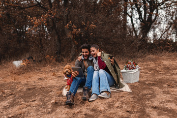 Couple at picnic sitting in blanket and using tablet. Next to them dog and basket with food. Autumn time.