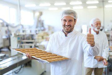 Young Caucasian worker in work wear holding tray with cookies and giving thumbs up while standing in food factory.