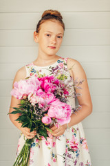 Summer portrait of adorable little girl wearing beautiful occasional dress, holding big peonies bouquet