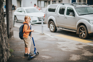 Little boy going to cross the road with car traffic, wearing school backpack, riding scooter