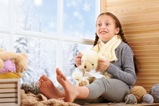 Child girl is sitting on a window sill and playing with bear toy. Beautiful view outside the window - sunny day in winter and snow.