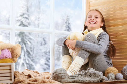 Child girl is sitting on a window sill and playing with bear toy. Beautiful view outside the window - sunny day in winter and snow.