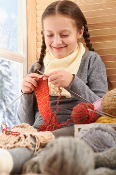 Child girl is sitting on a window sill with wool yarns and knitting. Beautiful view outside the window - sunny day in winter and snow.
