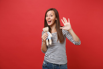 Portrait of funny young woman in striped clothes holding teddy bear plush toy showing palm, waving hand isolated on red wall background. People sincere emotions, lifestyle concept. Mock up copy space.