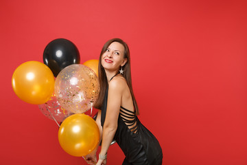 Fototapeta na wymiar Smiling young woman in little black dress celebrating looking up and holding air balloons isolated on bright red background. St. Valentine's Day, Happy New Year, birthday mockup holiday party concept.