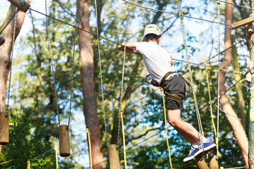 Naklejka na ściany i meble Cute boy enjoying activity in climbing adventure park at sunny summer day. Kid climbing in rope playground structure. Safe climbing with helmet insurance. Child in forest adventure park, extreme sport