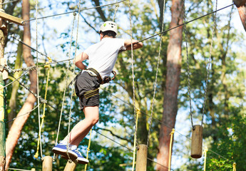 Naklejka na ściany i meble Cute boy enjoying activity in climbing adventure park at sunny summer day. Kid climbing in rope playground structure. Safe climbing with helmet insurance. Child in forest adventure park, extreme sport