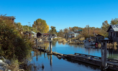 Fototapeta na wymiar An old fishermen village located in the slough of river. Old houses with moorings and boats near the shore. Early autumn, old grass and yellow trees against the blue sky. 
