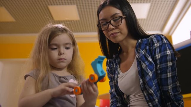 Positive female mixed race teacher helping cute little girl to make toy from colorful plastic construction blocks in kindergarten. Preschool teacher with child playing with didactic toys in class.