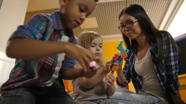 Portrait of beautiful teacher with multinational diverse kids playing with colorful didactic toys in kindergarten. Preschool multi ethnic children creating toys from constructing blocks in playroom.