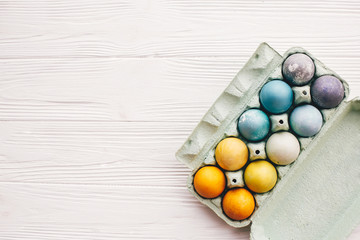 Stylish easter eggs in carton tray on white wooden background, top view. Modern colorful easter eggs painted with natural dye in different colors. Happy Easter, eco concept. Space for text