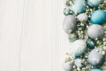 Stylish easter eggs with spring flowers on white wooden background with space for text, flat lay. Modern easter eggs painted with natural dye in blue and grey color. Happy Easter